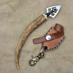 May include: A handmade hunting arrowhead with a deer antler handle and a silver blade. The arrowhead is displayed next to a brown leather sheath with a metal clasp and key ring. The background is a textured, tan surface.