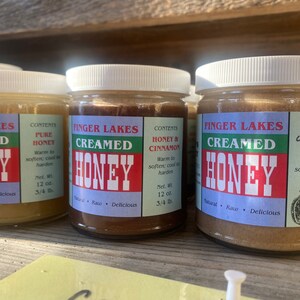 May include: Three glass jars of Finger Lakes creamed honey. The jars are labeled with the contents: Pure Honey, Honey & Cinnamon, and Honey Cinnamon Nutmeg Ginger. Each jar has a white lid and a green label with white text.