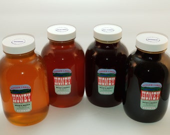 Puede incluir: Cuatro tarros de vidrio de miel, cada uno con una tapa blanca y una etiqueta roja y blanca que dice "Finger Lakes Honey, Waid's Honey". Los tarros están dispuestos en una fila sobre una superficie blanca. La miel en los tarros es de varios colores, incluyendo ámbar claro, ámbar oscuro y marrón oscuro.