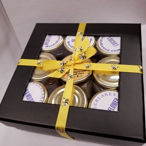 May include: A black gift box with a clear window, filled with eight small glass jars of honey. The jars are tied with a yellow ribbon with a bee design. The jars are labeled with different types of honey, including Wildflower, Bamboo, Gingerlake, and Locust.