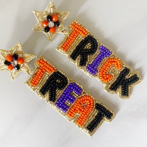 May include: A pair of beaded earrings with gold star charms. The earrings spell out "Trick" and "Treat" in black, orange, and purple beads.
