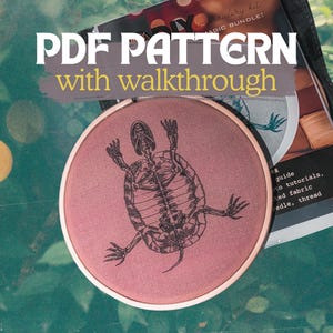 Peut inclure: Un tambour à broder circulaire contenant un tissu rose poudré avec le dessin au trait noir d'un squelette de tortue. Le texte PDF PATTERN with walkthrough est affiché au-dessus du tambour, posé sur un guide d'instructions imprimé.