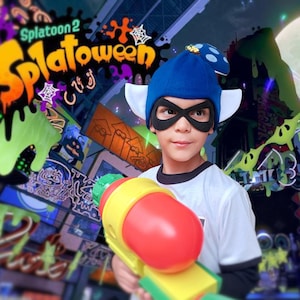 May include: A person wearing a blue hat with white ear-like accents and a black mask, holding a red and yellow toy gun. The image features the text "Splatoon 2 Splatoween" in orange and green.