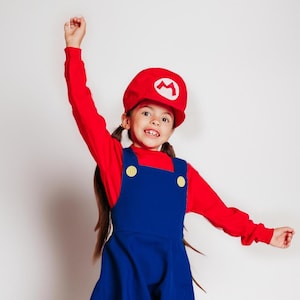May include: A child wearing a red long-sleeve shirt and a blue overall dress. The child is also wearing a red hat with a white "M" symbol. The outfit is a costume, likely inspired by a video game character.