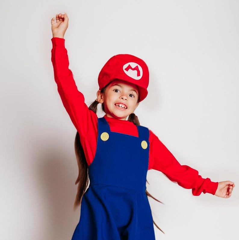 May include: A child wearing a red long-sleeve shirt and a blue overall dress. The child is also wearing a red hat with a white "M" symbol. The outfit is a costume, likely inspired by a video game character.