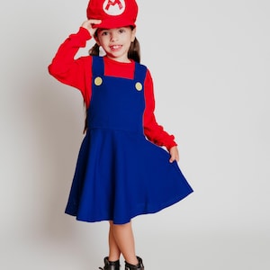 May include: A child in a red long-sleeve shirt and a blue dress with yellow buttons, wearing a red hat with a white "M" logo. The child is also wearing black boots. The outfit resembles a popular video game character.