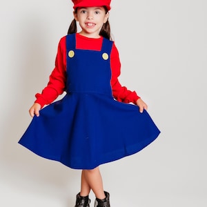 May include: A child's costume inspired by a video game character. The outfit includes a red cap with a white emblem, a red long-sleeved shirt, and a blue dress with yellow buttons. The child is also wearing black boots.