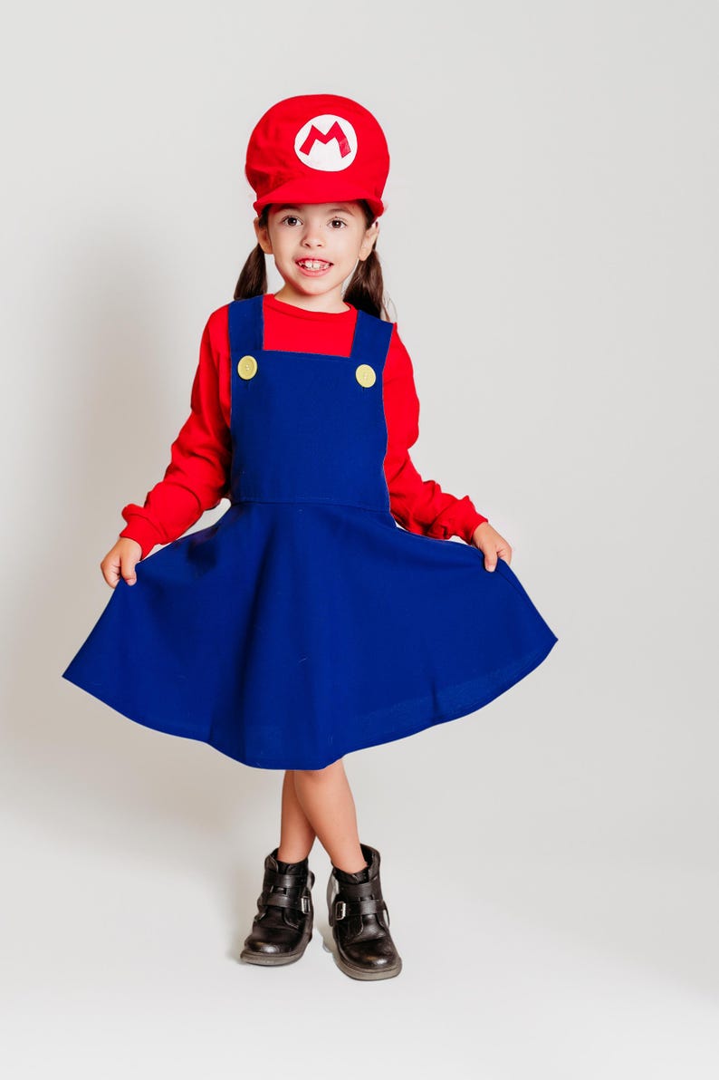 May include: A child's costume inspired by a video game character. The outfit includes a red cap with a white emblem, a red long-sleeved shirt, and a blue dress with yellow buttons. The child is also wearing black boots.
