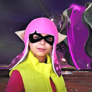 May include: A bright pink squid-shaped hat with long side flaps, featuring a black eye mask, paired with a vibrant yellow sleeveless vest over a pink long-sleeved shirt. The hat has light-colored pointed ears and a dark pink gradient pattern.