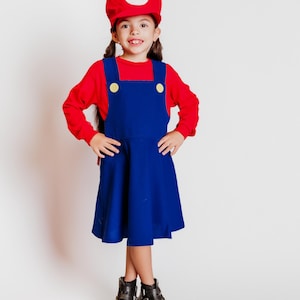 May include: A child dressed in a Mario costume, featuring a red hat with a white 'M' emblem, a red long-sleeve shirt, and a blue overall dress with yellow buttons. The outfit is completed with black boots.