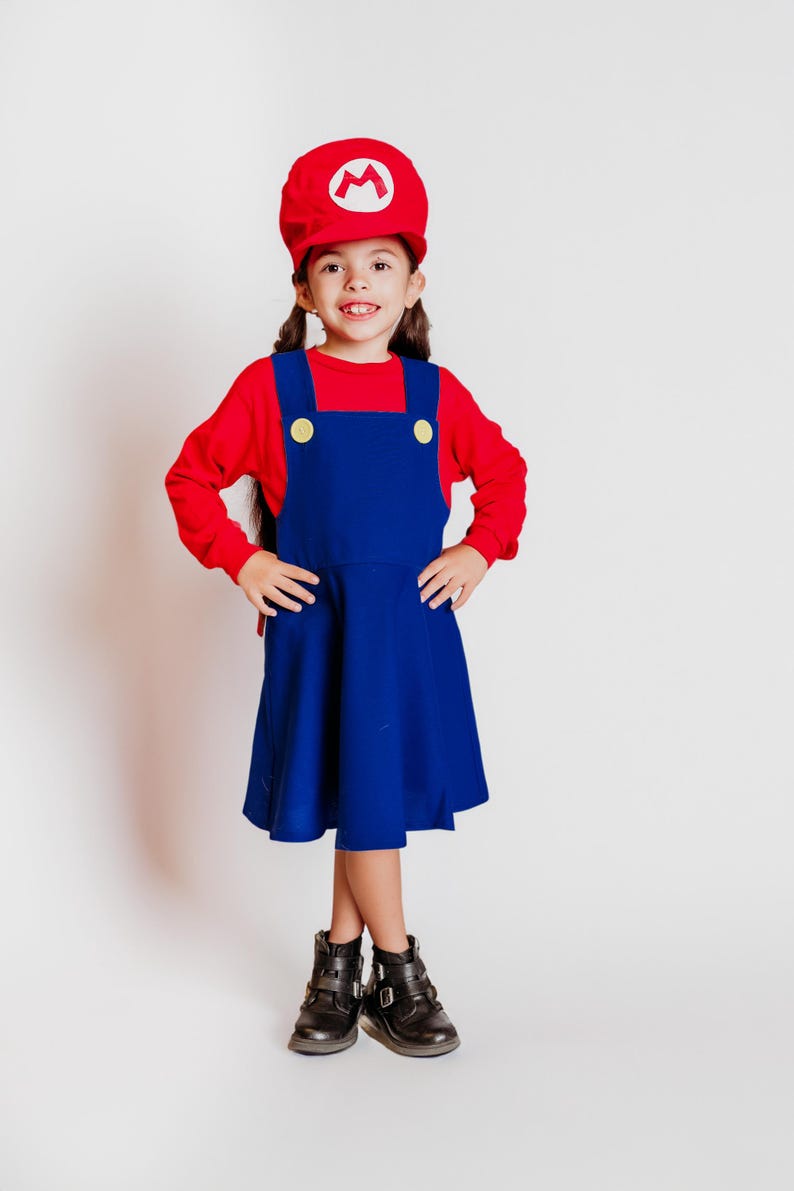May include: A child dressed in a Mario costume, featuring a red hat with a white 'M' emblem, a red long-sleeve shirt, and a blue overall dress with yellow buttons. The outfit is completed with black boots.