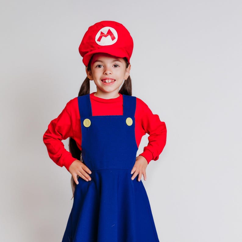 May include: A child wearing a red long-sleeved shirt and a blue overall dress. The outfit includes a red cap with a white emblem. The overall dress has yellow buttons. The child is smiling.