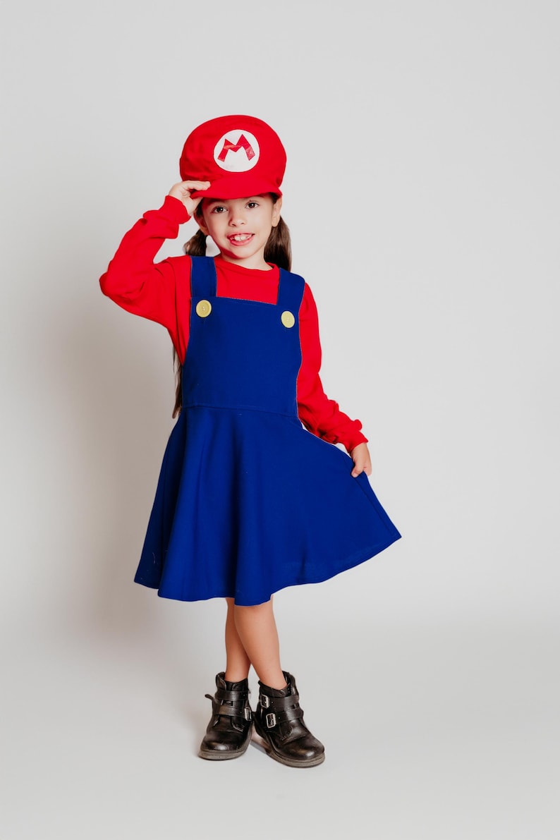 May include: A child in a red long-sleeved shirt and a blue dress, styled as a Mario costume. The outfit includes a red hat with a white "M" emblem and black boots. The dress has yellow button details.