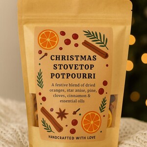 Könnte beinhalten: Ein Kraftpapierbeutel mit der Aufschrift "CHRISTMAS STOVETOP POTPOURRI" und Illustrationen von Orangen, Zimtstangen und Sternanis. Der Text beschreibt eine festliche Mischung aus getrockneten Orangen, Sternanis, Kiefer, Nelken, Zimt und ätherischen Ölen. "HANDCRAFTED WITH LOVE" ist ebenfalls aufgedruckt.