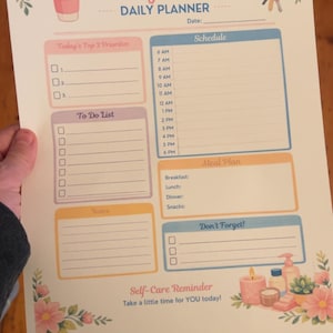 May include: A printed daily planner titled "Busy Mom" with sections for priorities, to-do list, schedule, meal plan, notes, and reminders. The planner features floral and coffee cup illustrations, with the text "Self-Care Reminder" at the bottom.