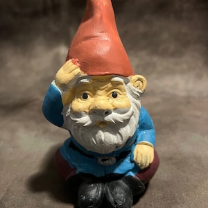 May include: A small, seated garden gnome figurine with a red hat, blue clothing, and a white beard. The gnome is looking forward with a serious expression.