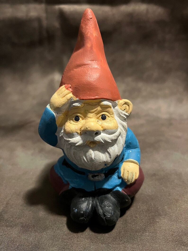 May include: A small, seated garden gnome figurine with a red hat, blue clothing, and a white beard. The gnome is looking forward with a serious expression.