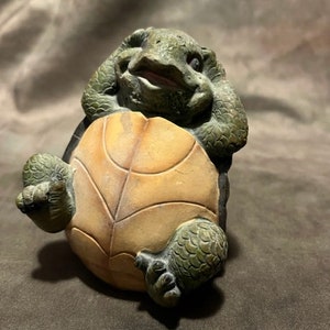 May include: A small, green and brown ceramic turtle figurine with its head resting on its shell. The turtle has a playful expression on its face.