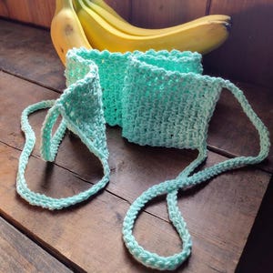 May include: A light blue crocheted bag with a long strap. The bag is lying on a wooden surface with a bunch of bananas behind it.