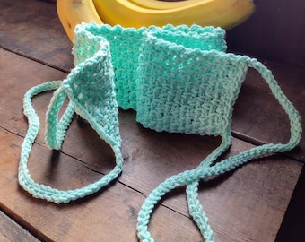 May include: A light blue crocheted bag with a long strap. The bag is lying on a wooden surface with a bunch of bananas behind it.