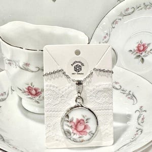 May include: A silver chain necklace with a round pendant featuring a pink rose on a white background. The pendant is framed with a silver border. The necklace is displayed on a white card with the text "Anthology Art House".