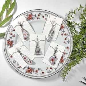 May include: Five necklaces with floral patterned porcelain pendants on white cards with a silver chain. The necklaces are displayed on a white plate with a floral pattern.