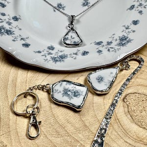 May include: Three silver charm necklaces with blue and white porcelain accents. The charms are shaped like a teardrop, a rectangle, and a bookmark. The charms are all set in silver and have a delicate floral design.
