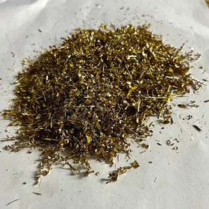 May include: A pile of gold-colored metal shavings on a white surface.