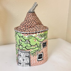 May include: A ceramic jar shaped like a small house with a removable conical roof lid. The structure features a red brick pattern, a white door with black vertical lines, two black-framed windows, and green painted foliage climbing the walls.