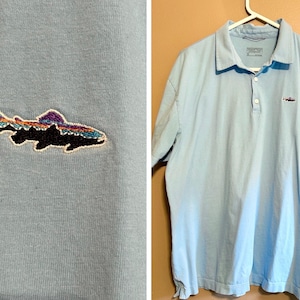 May include: Light blue polo shirt with a small embroidered fish design on the chest. The fish is black with colorful accents of purple, orange, and blue. The shirt is hanging on a white hanger.