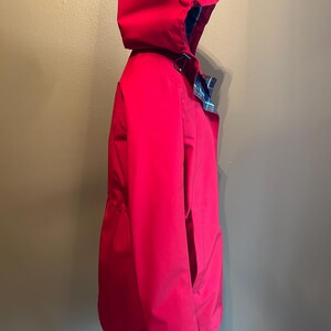 Puede incluir: Una chaqueta con capucha de color rojo brillante con cintura ajustable con cord&oacute;n y forro a cuadros visible en el borde de la capucha. La chaqueta tiene un dise&ntilde;o cl&aacute;sico, con un bolsillo lateral visible. La chaqueta es de un material liso, posiblemente resistente al agua.