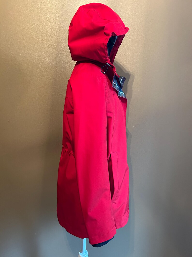 Puede incluir: Una chaqueta con capucha de color rojo brillante con cintura ajustable con cord&oacute;n y forro a cuadros visible en el borde de la capucha. La chaqueta tiene un dise&ntilde;o cl&aacute;sico, con un bolsillo lateral visible. La chaqueta es de un material liso, posiblemente resistente al agua.