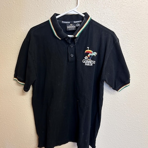 May include: Black polo shirt with a green, white, and orange striped collar and sleeve cuffs. The shirt features the Guinness logo and the words "Guinness Dublin" embroidered on the chest.