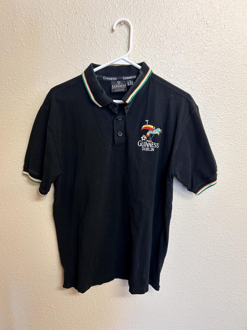 May include: Black polo shirt with a green, white, and orange striped collar and sleeve cuffs. The shirt features the Guinness logo and the words "Guinness Dublin" embroidered on the chest.
