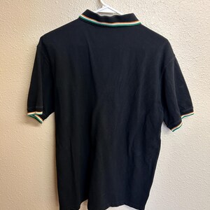 May include: Black polo shirt with a green, white, and orange striped collar and sleeve cuffs. The shirt is hanging on a white hanger against a neutral wall. The shirt is made of a soft material.