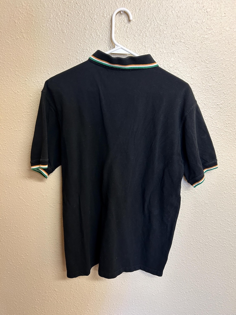 May include: Black polo shirt with a green, white, and orange striped collar and sleeve cuffs. The shirt is hanging on a white hanger against a neutral wall. The shirt is made of a soft material.