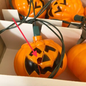 May include: A string of orange jack-o'-lantern lights with black facial features. The lights are connected by green wires and are in a cardboard box. One pumpkin has a crack in it.