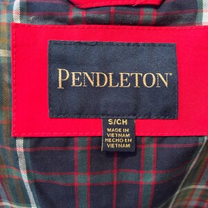 Puede incluir: Primer plano de una etiqueta de prenda con el nombre de la marca "PENDLETON" en letras doradas sobre un fondo azul marino. La etiqueta est&aacute; colocada sobre una tela roja, con un estampado de cuadros visible en el fondo. La etiqueta tambi&eacute;n incluye la talla "S/CH" y el texto "MADE IN VIETNAM HECHO EN VIETNAM".
