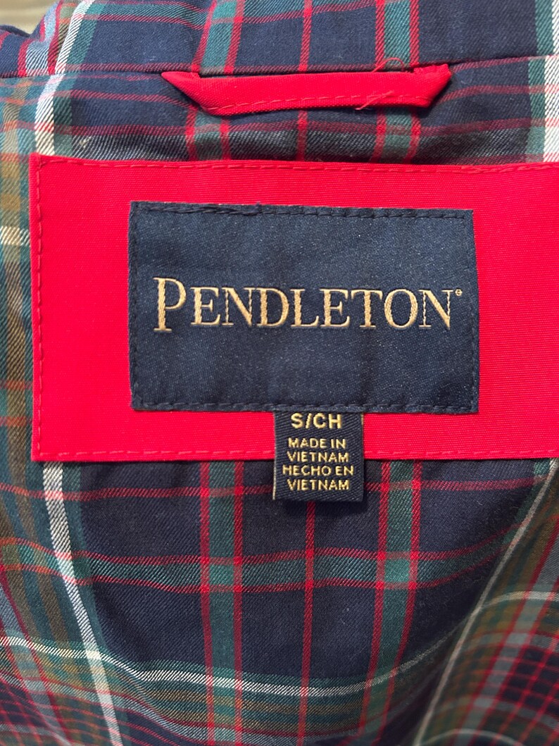 Puede incluir: Primer plano de una etiqueta de prenda con el nombre de la marca "PENDLETON" en letras doradas sobre un fondo azul marino. La etiqueta est&aacute; colocada sobre una tela roja, con un estampado de cuadros visible en el fondo. La etiqueta tambi&eacute;n incluye la talla "S/CH" y el texto "MADE IN VIETNAM HECHO EN VIETNAM".