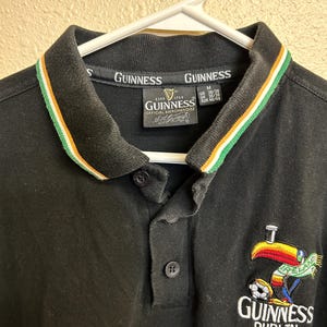 May include: Black polo shirt with a collar featuring green, white, and orange stripes. The shirt has a Guinness logo and a colorful toucan graphic with the word "Ireland" and a soccer ball. The shirt is hanging on a white hanger.
