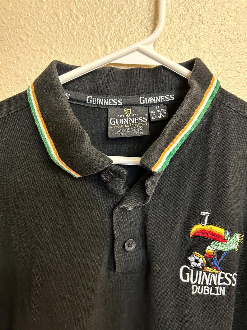 May include: Black polo shirt with a collar featuring green, white, and orange stripes. The shirt has a Guinness logo and a colorful toucan graphic with the word "Ireland" and a soccer ball. The shirt is hanging on a white hanger.
