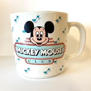Pode incluir: Caneca de cer&acirc;mica branca com um design do Mickey Mouse Club. A caneca apresenta uma imagem de desenho animado do Mickey Mouse com notas musicais no fundo. O texto "Mickey Mouse Club" est&aacute; impresso em vermelho e azul na caneca.