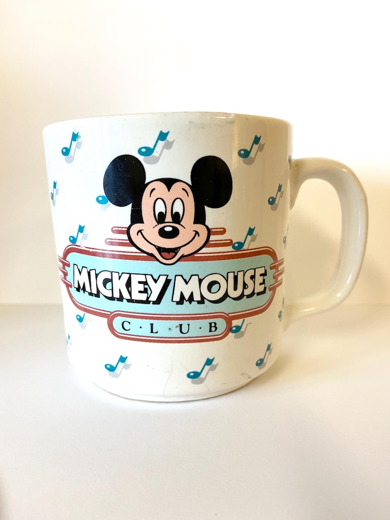 Pode incluir: Caneca de cer&acirc;mica branca com um design do Mickey Mouse Club. A caneca apresenta uma imagem de desenho animado do Mickey Mouse com notas musicais no fundo. O texto "Mickey Mouse Club" est&aacute; impresso em vermelho e azul na caneca.