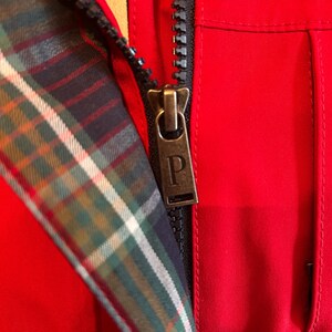 Puede incluir: Primer plano de una chaqueta roja con cremallera negra y forro a cuadros. El tirador de la cremallera es de color bronce con la letra "P". El forro a cuadros presenta colores verdes, azules y blancos.