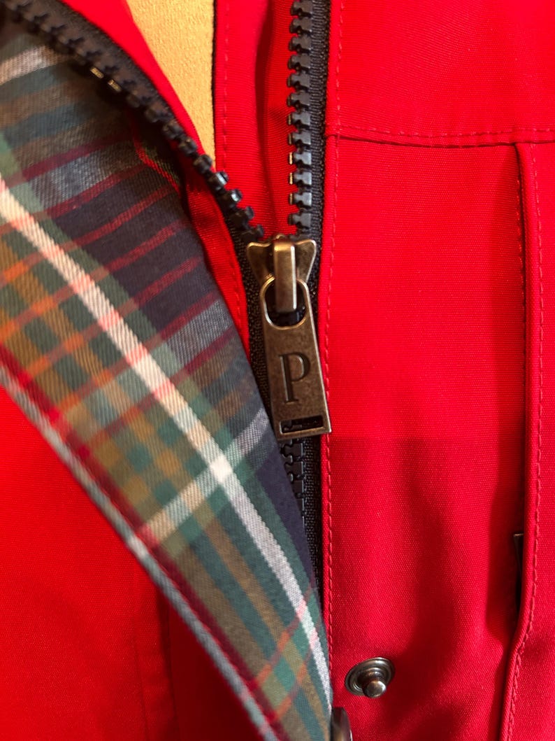 Puede incluir: Primer plano de una chaqueta roja con cremallera negra y forro a cuadros. El tirador de la cremallera es de color bronce con la letra "P". El forro a cuadros presenta colores verdes, azules y blancos.