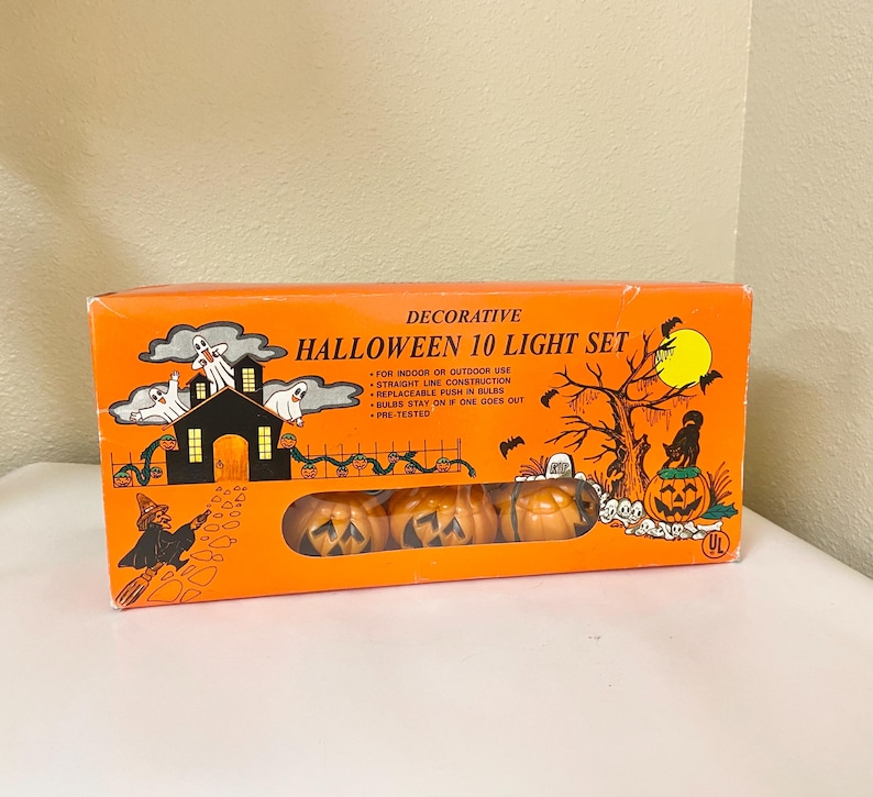 May include: An orange box with the text "DECORATIVE HALLOWEEN 10 LIGHT SET" features a Halloween-themed illustration. The illustration includes a haunted house, ghosts, a witch, and jack-o'-lantern lights. The box contains pumpkin-shaped lights.