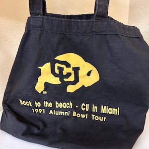 May include: A black canvas tote bag with two shoulder straps. The front features a yellow graphic of a buffalo silhouette with the letters CU inside, and the text: Back to the beach - CU in Miami 1991 Alumni Bowl Tour printed in yellow.