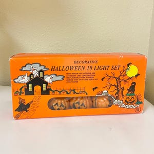 May include: Decorative Halloween 10 light set in an orange box. The box features illustrations of a haunted house, ghosts, a witch, and jack-o'-lanterns. The set includes pumpkin-shaped lights.