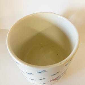 Pode incluir: Caneca de cer&acirc;mica branca com um padr&atilde;o de peixes e ondas estilizados em azul e vermelho. A caneca est&aacute; vazia.