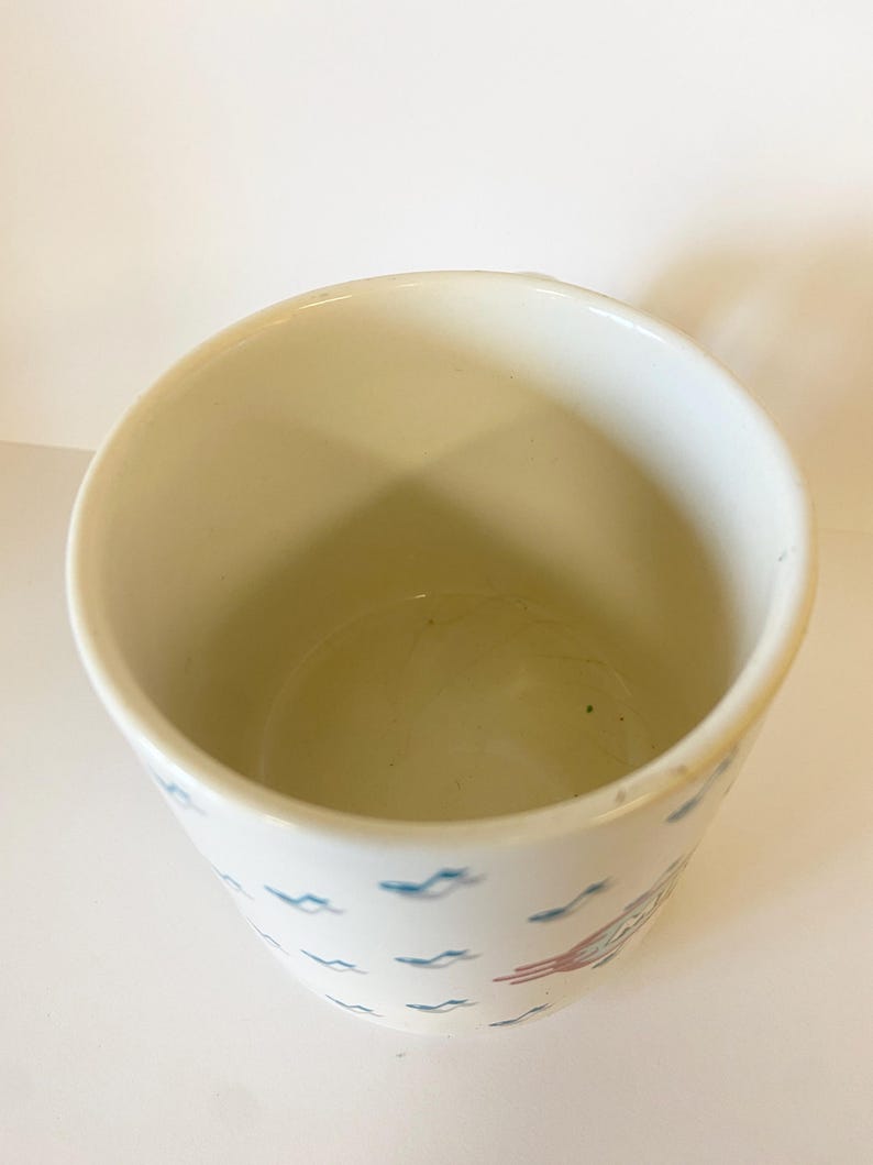 Pode incluir: Caneca de cer&acirc;mica branca com um padr&atilde;o de peixes e ondas estilizados em azul e vermelho. A caneca est&aacute; vazia.